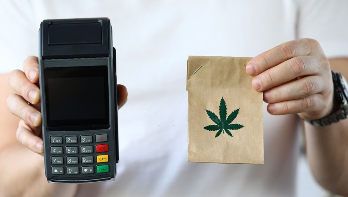 Visa Closes a Loophole That Allows Buying Weed With a Bank Card: A man holding a POS terminal in one hand and a brown paper pouch with a weed logo on it in the other